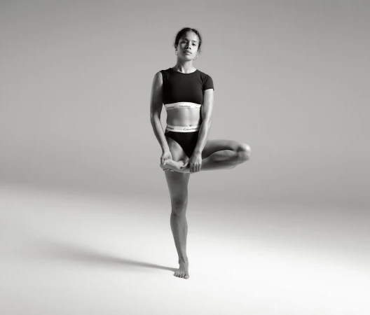 A woman in athletic wear balances in a tree pose against a neutral backdrop, highlighting fitness and minimalist sportswear fashion.