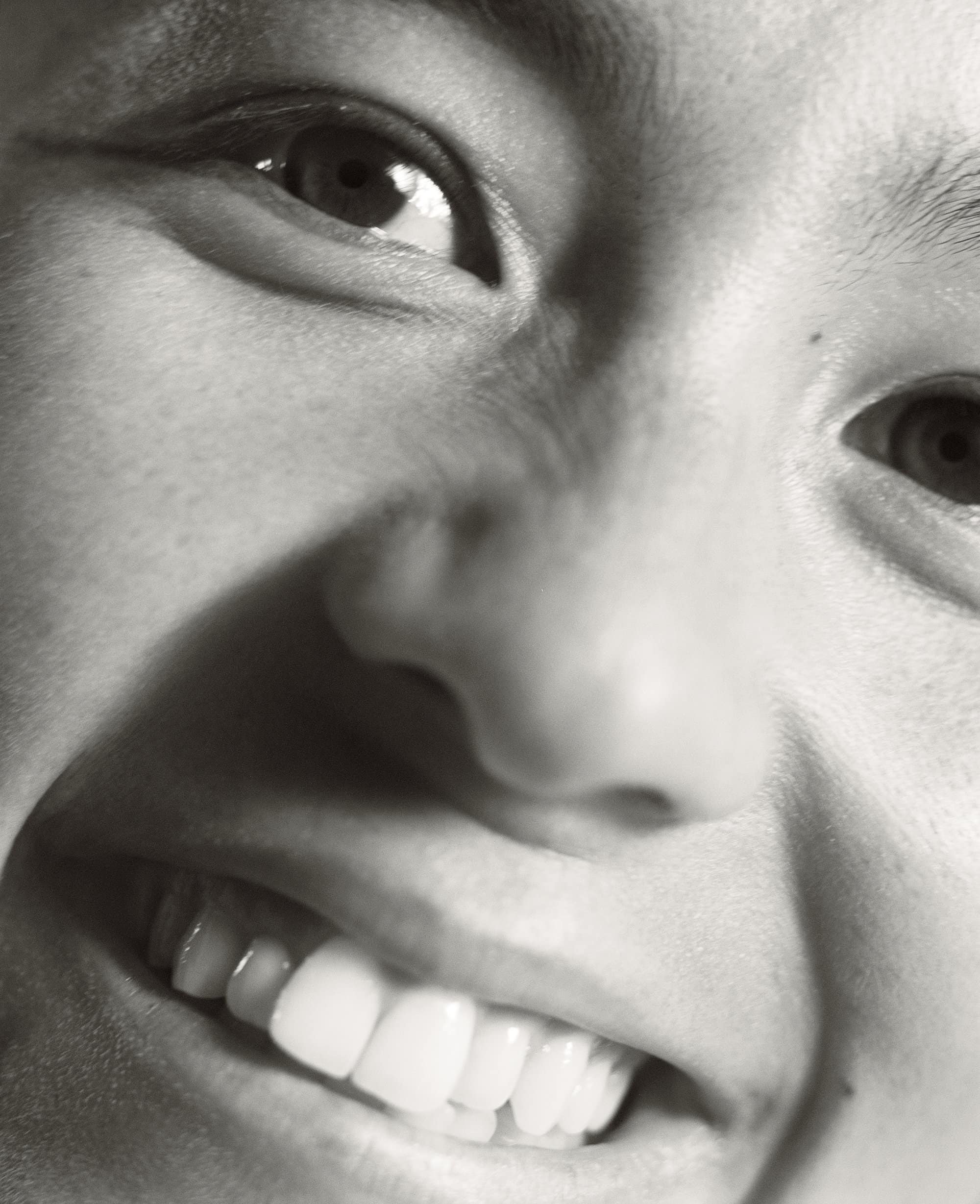 Close-up black and white portrait of a smiling woman. A high-angle captures her face, highlighting her eyes, nose, mouth, and teeth.