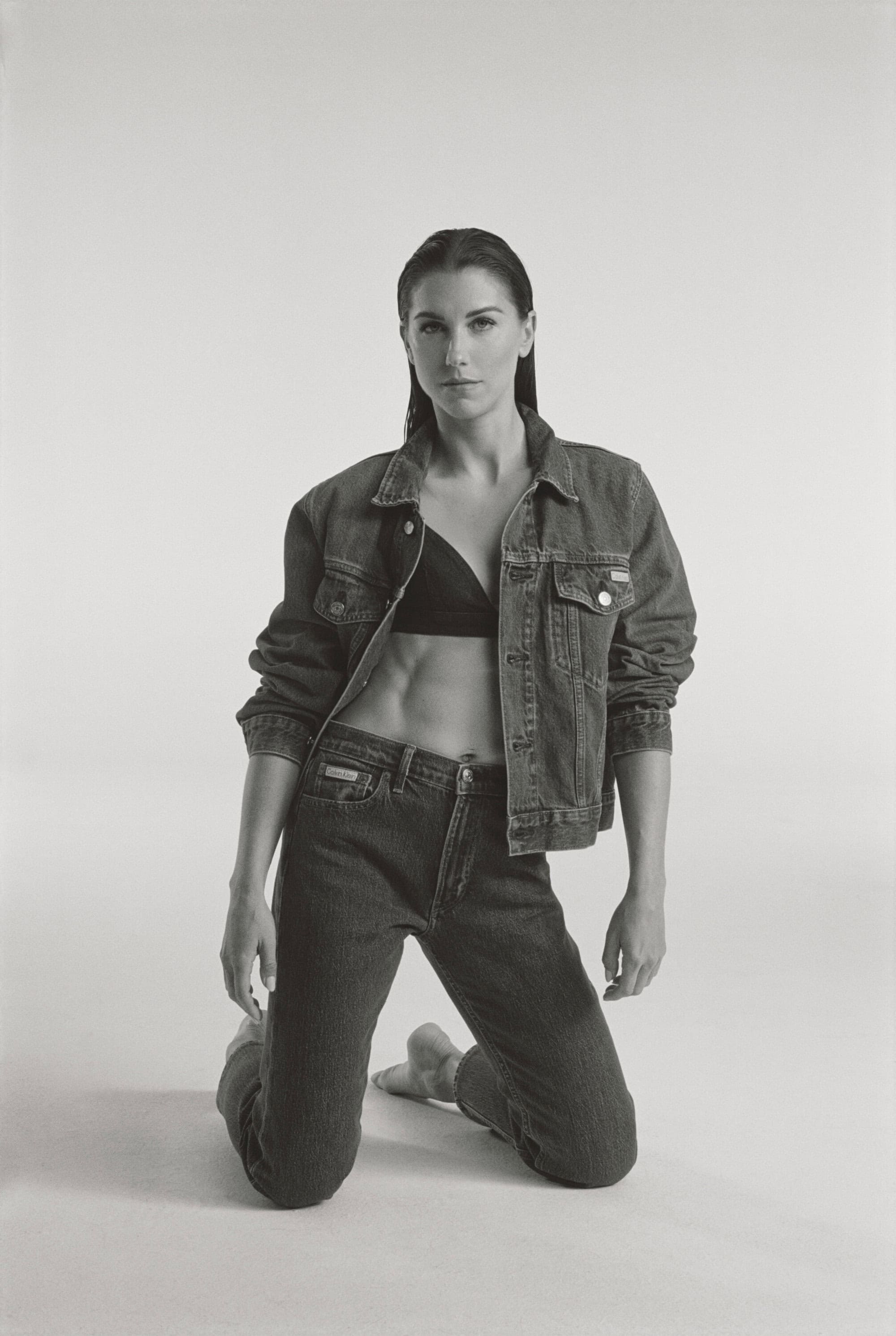 A woman with athletic physique kneels wearing a denim jacket and jeans, looking at the camera in a studio environment with soft lighting.
