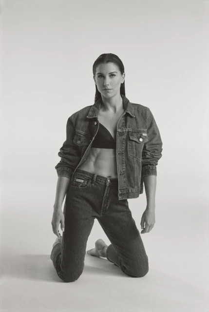 A woman with athletic physique kneels wearing a denim jacket and jeans, looking at the camera in a studio environment with soft lighting.