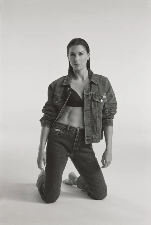 A woman with athletic physique kneels wearing a denim jacket and jeans, looking at the camera in a studio environment with soft lighting.