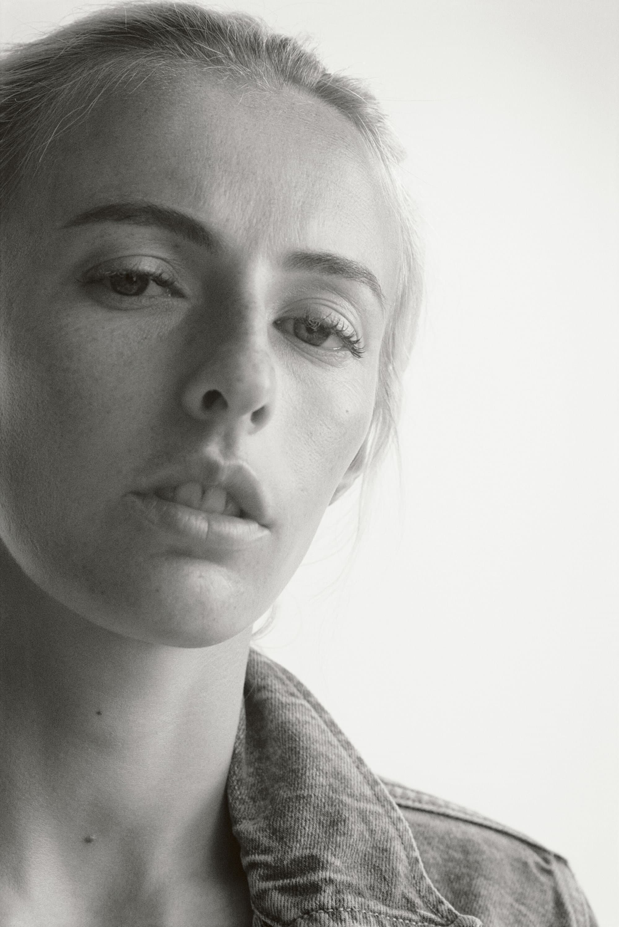 Monochrome portrait of a young adult woman with fair skin wearing a denim jacket, captured in a studio setting with diffused lighting.