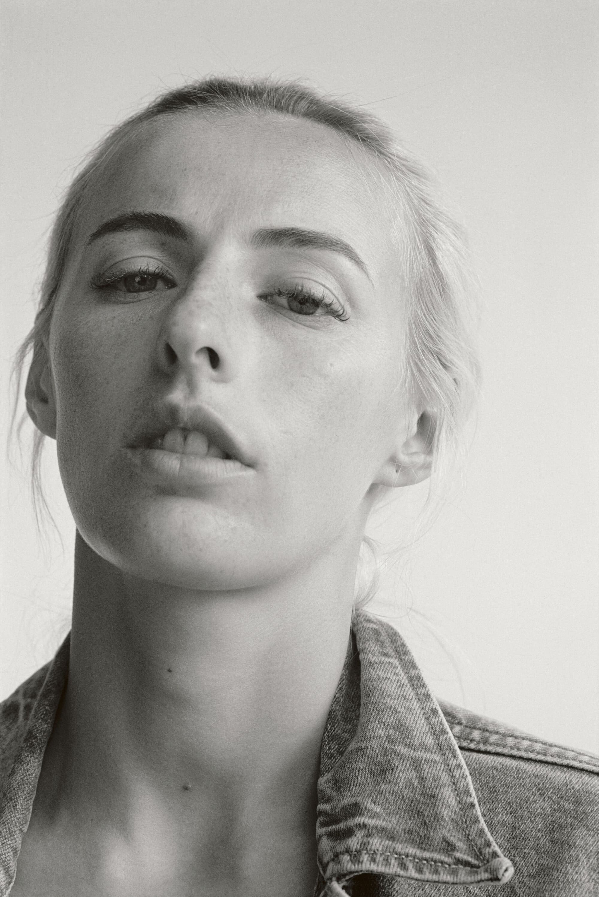 Monochrome portrait of a fair-skinned young adult woman looking at the camera with a denim jacket, against a white background.
