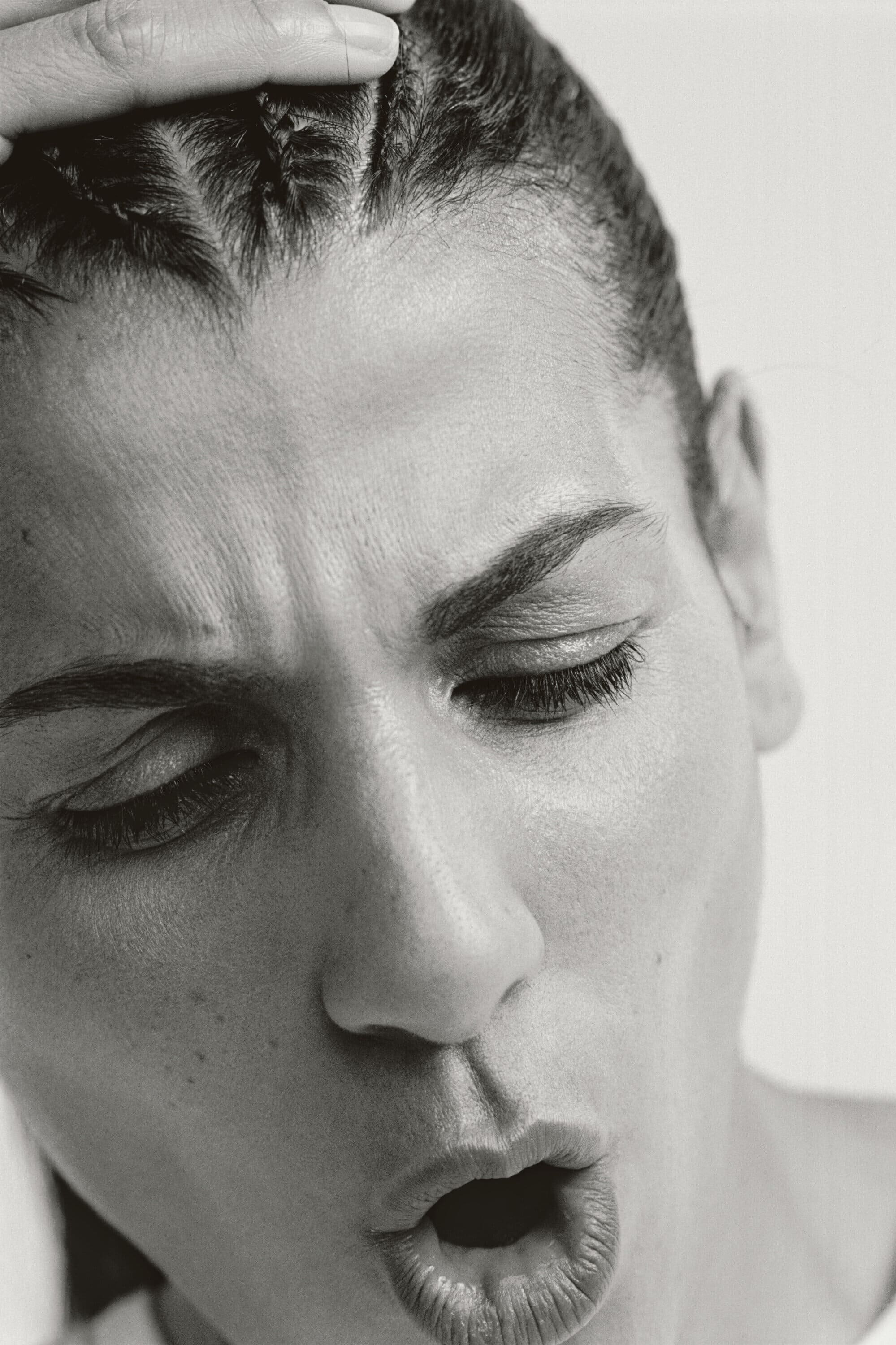 Monochrome close-up of an adult woman with braided hair touching her forehead, her eyes closed and her mouth open.