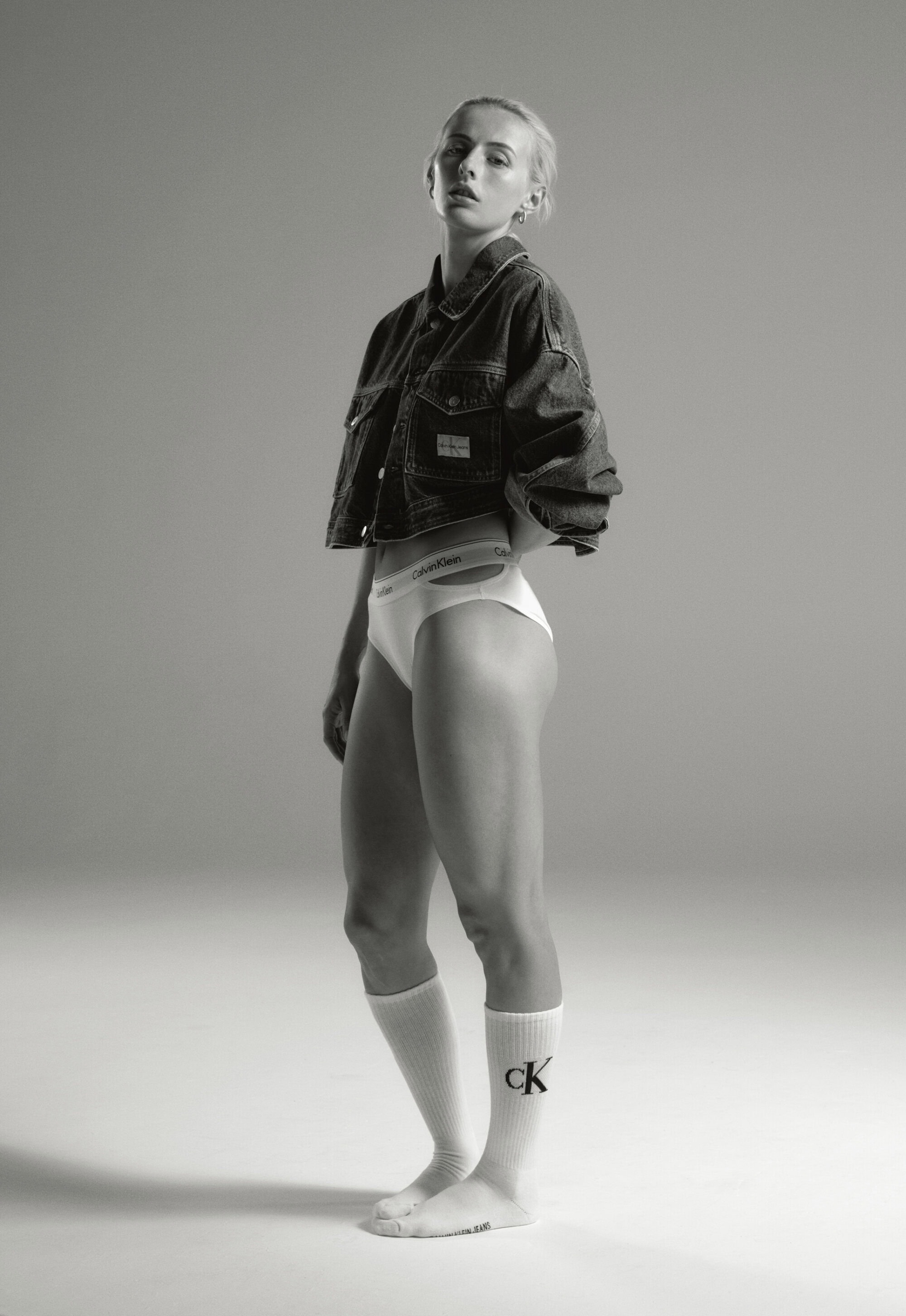 Blonde woman in a denim jacket and underwear poses in a studio. The model wears knee-high socks with the CK logo.