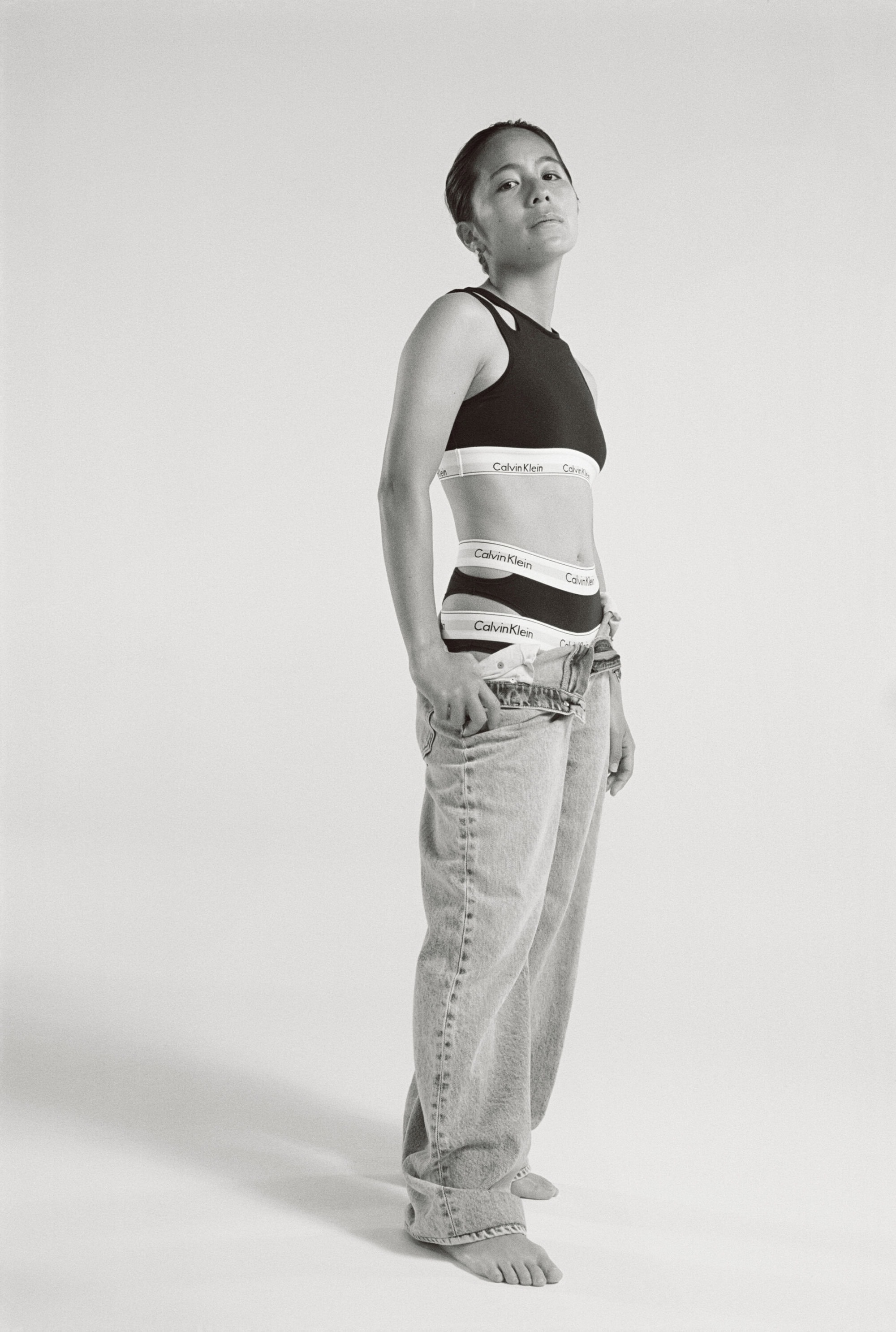 Monochromatic studio shot of a woman in Calvin Klein underwear and jeans, striking a confident pose against a bright backdrop.