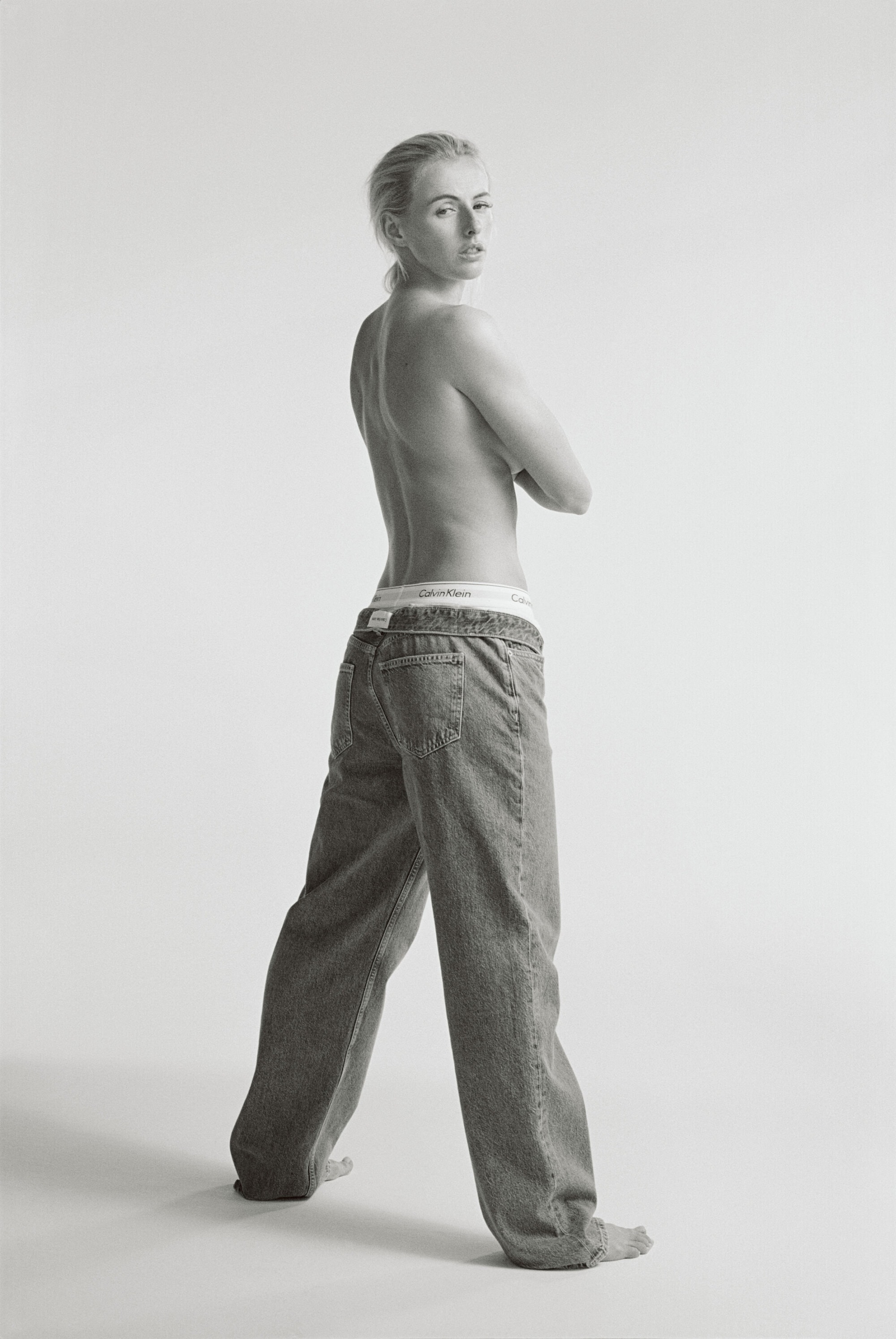 Full figure studio shot of adult woman with arms crossed, wearing jeans and visible underwear band, looking over her shoulder.