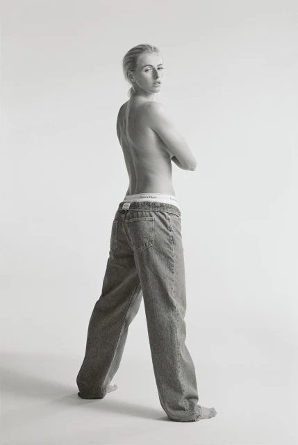 Full figure studio shot of adult woman with arms crossed, wearing jeans and visible underwear band, looking over her shoulder.