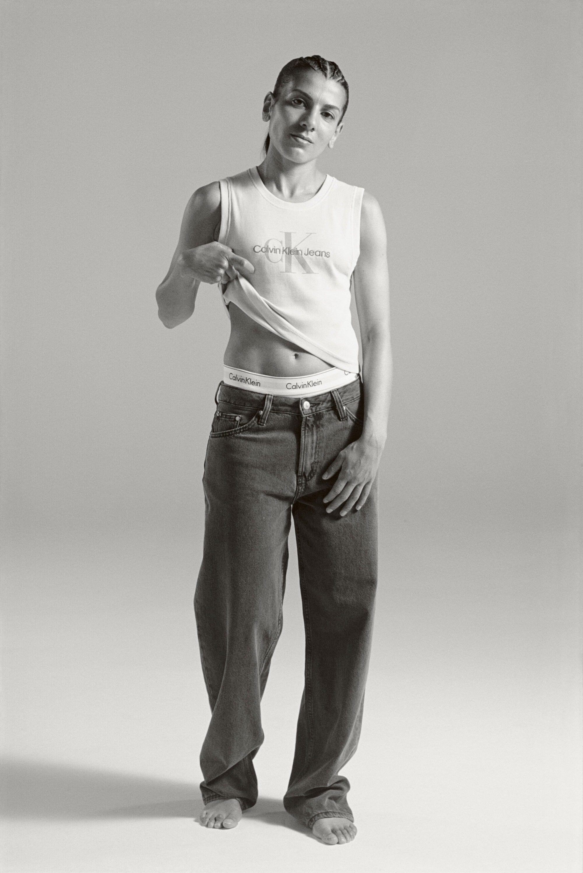 A woman poses in a Calvin Klein top and jeans, lifting her shirt to reveal the waistband, photographed in a studio setting.