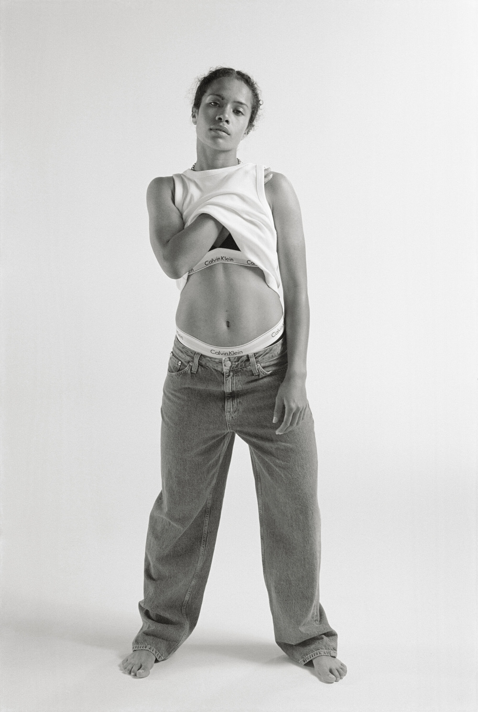 A woman stands in a photo studio, wearing a white tank top, blue jeans, and white underwear, looking directly at the camera.