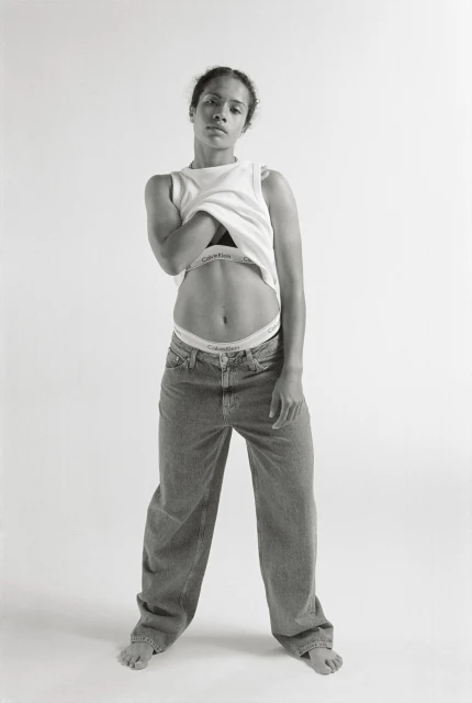 A woman stands in a photo studio, wearing a white tank top, blue jeans, and white underwear, looking directly at the camera.