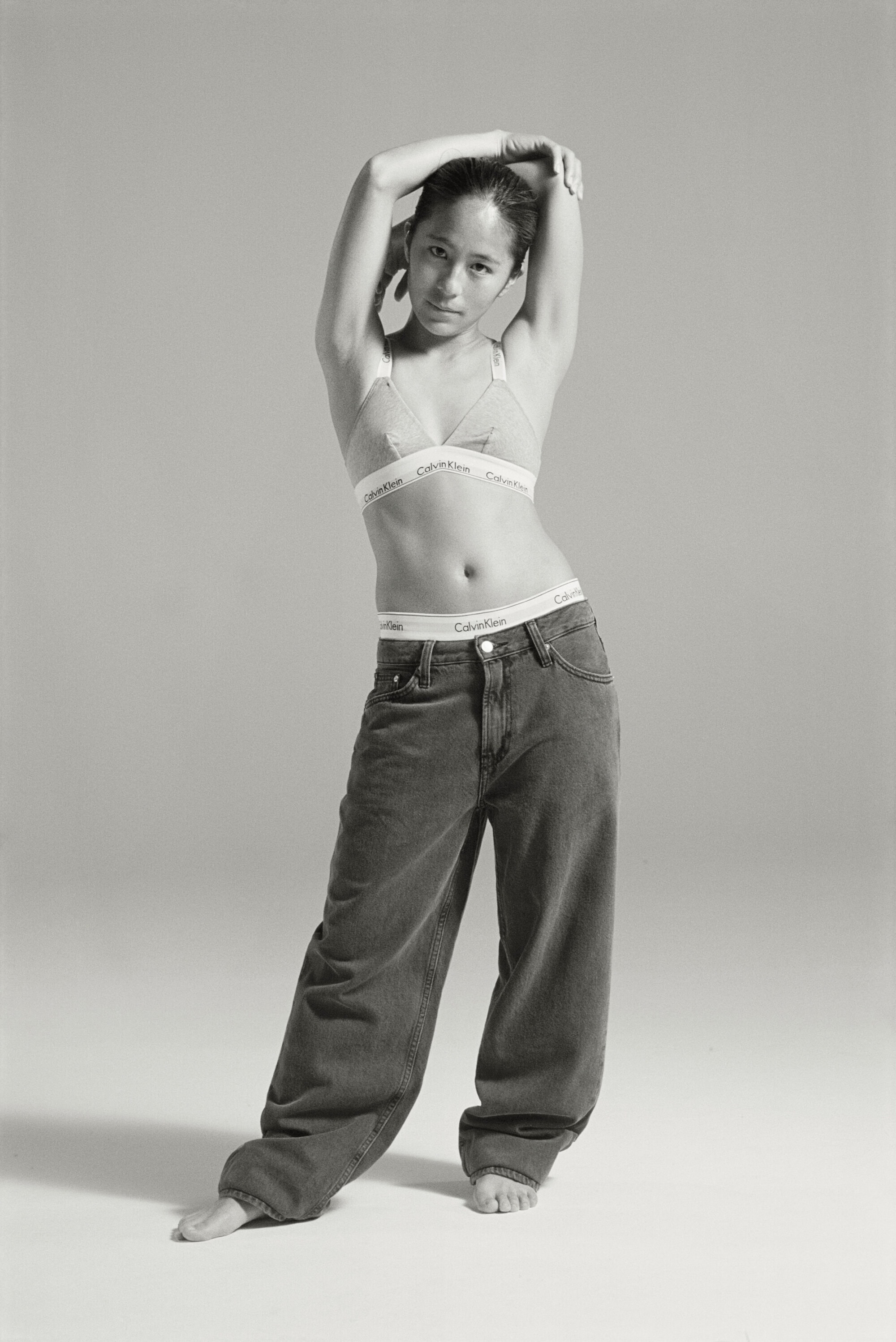 A slender, fair-skinned adult woman in jeans and a bra poses in a full shot against a neutral backdrop in a fashion forward style.