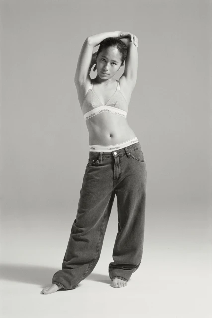 A slender, fair-skinned adult woman in jeans and a bra poses in a full shot against a neutral backdrop in a fashion forward style.