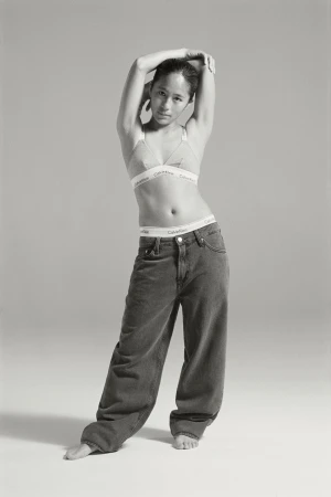 A slender, fair-skinned adult woman in jeans and a bra poses in a full shot against a neutral backdrop in a fashion forward style.