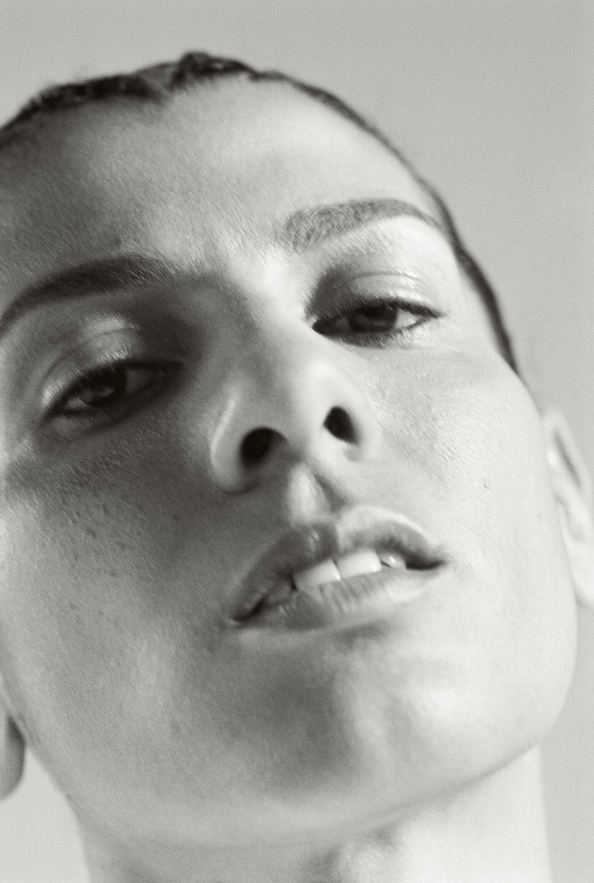 Close-up portrait of an adult female with slicked back dark hair and neutral expression, set against a plain light gray background.