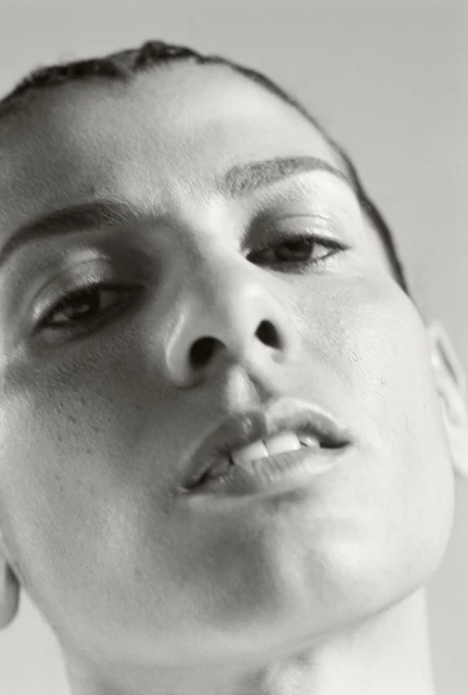 Close-up portrait of an adult female with slicked back dark hair and neutral expression, set against a plain light gray background.