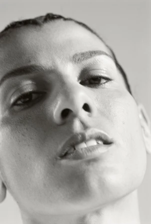 Close-up portrait of an adult female with slicked back dark hair and neutral expression, set against a plain light gray background.