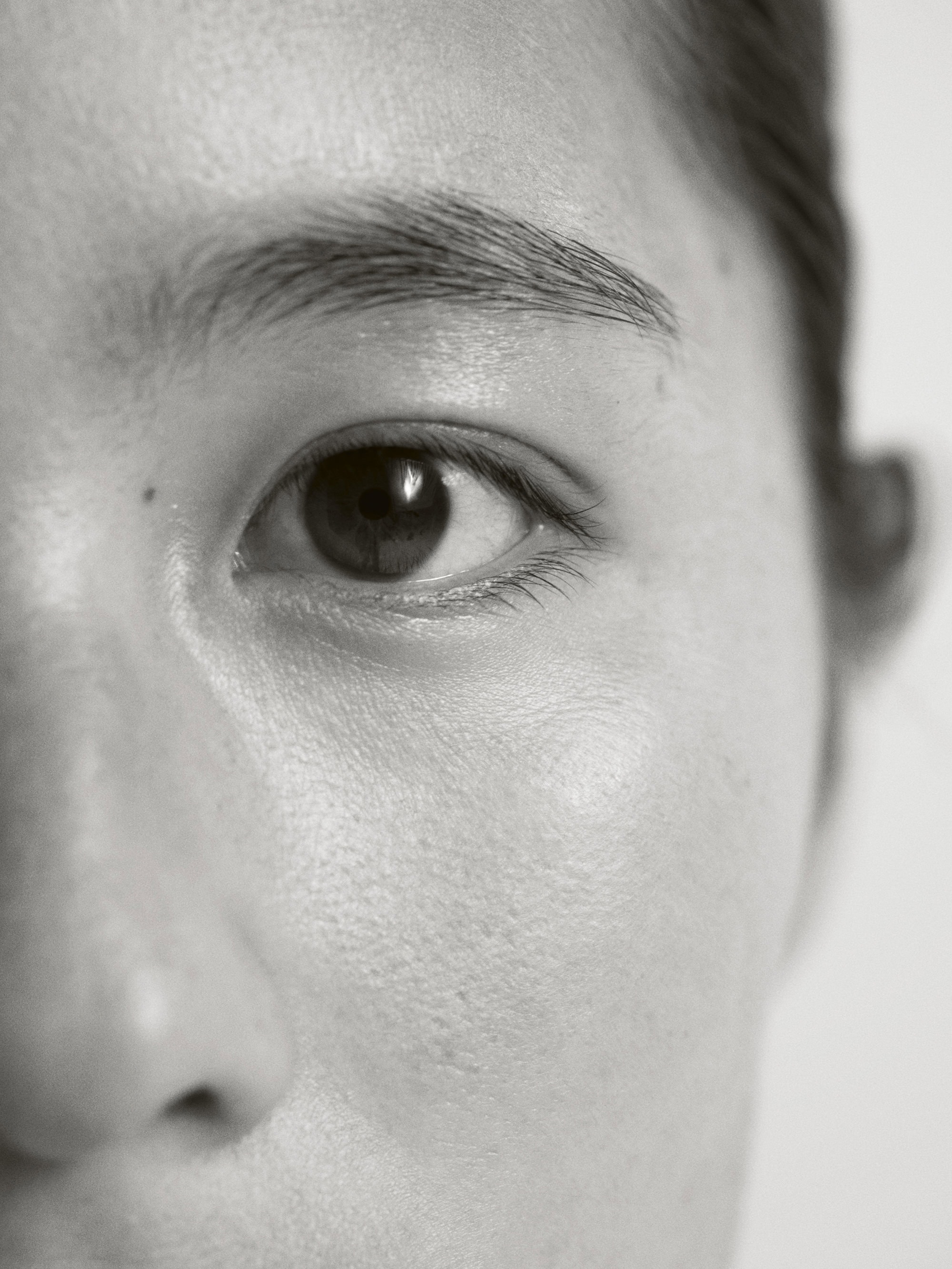 Close-up of an Asian woman's face, focusing on her eye, eyebrow, and skin texture. The image is in black and white with soft lighting.