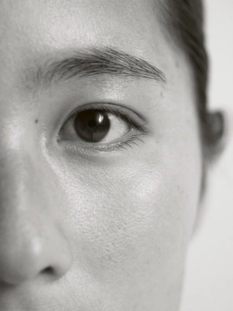Close-up of an Asian woman's face, focusing on her eye, eyebrow, and skin texture. The image is in black and white with soft lighting.