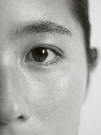 Close-up of an Asian woman's face, focusing on her eye, eyebrow, and skin texture. The image is in black and white with soft lighting.
