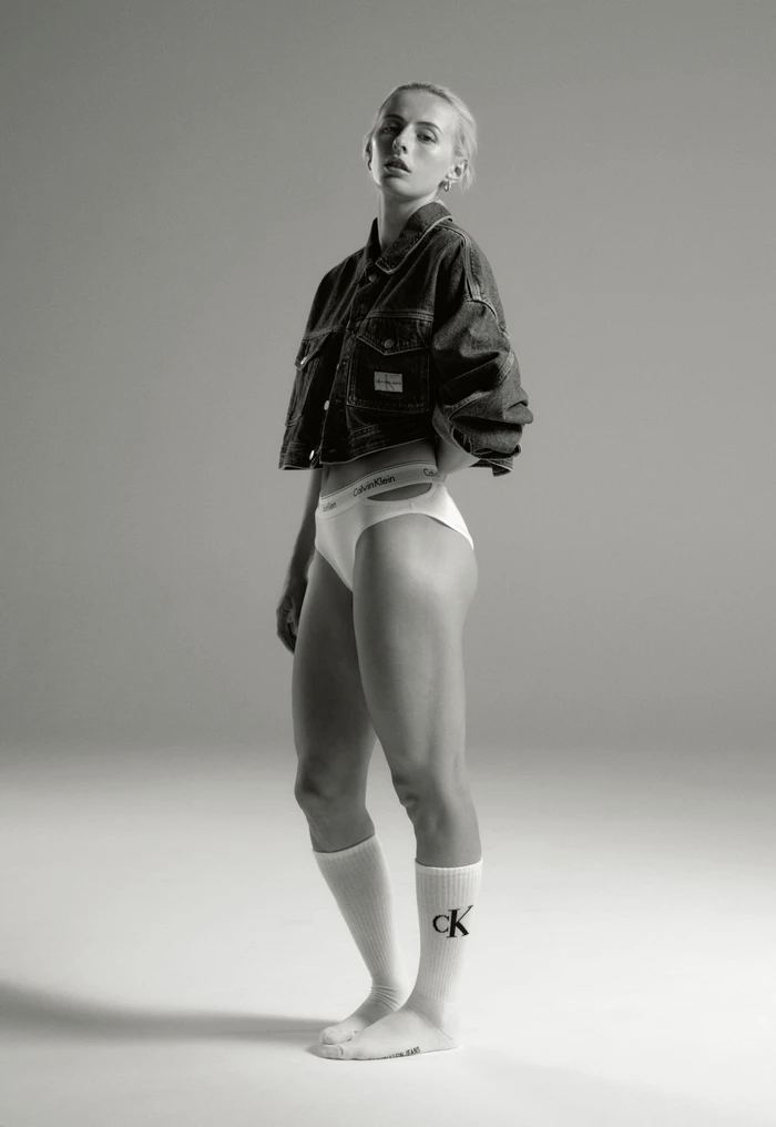 Model in denim jacket and white underwear standing against a plain grey studio backdrop