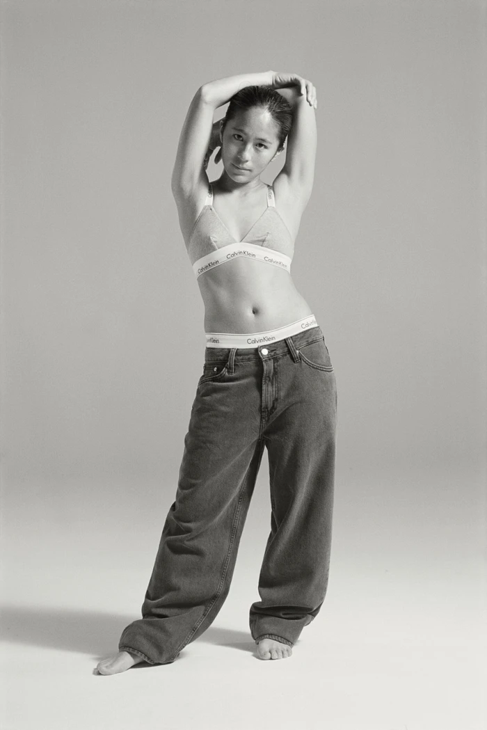 Woman in Calvin Klein bralette and relaxed jeans posing against a seamless grey backdrop.