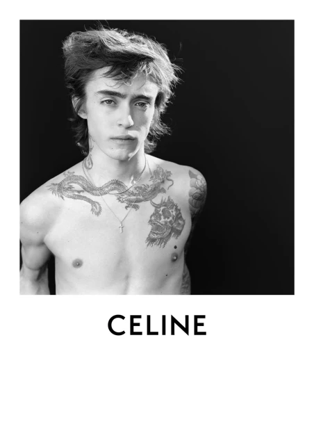 Black and white portrait of a young man with tattoos, wearing a silver cross necklace, set against a dark backdrop for a stark and stylish look.