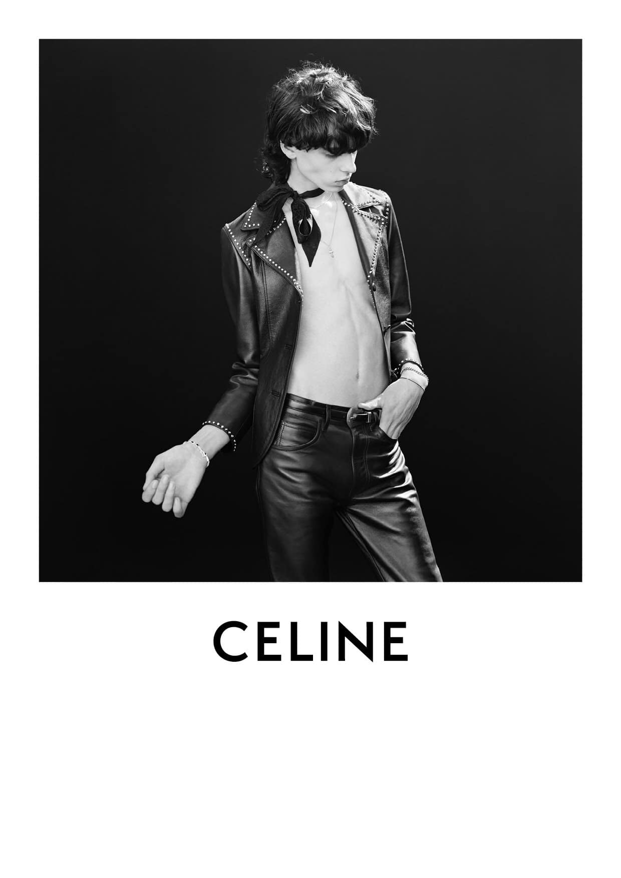 A person posing in a black leather jacket and black pants against a dark background, accessorized with a bracelet and neck scarf.