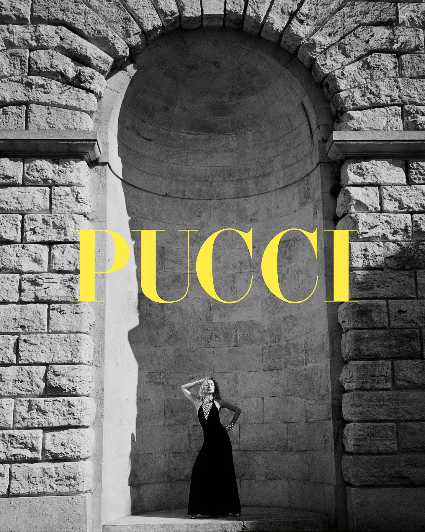 A woman in a black gown poses within a stone archway under bright, natural lighting, with the word 'PUCCI' emblazoned across the top.