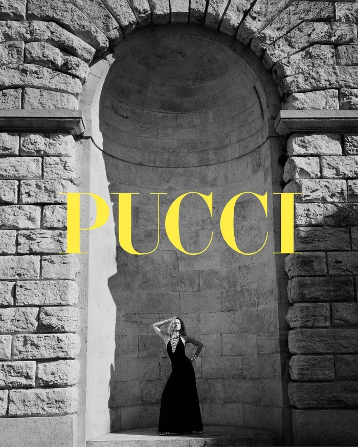 Model posing inside a grand stone arched alcove wearing a dark halter dress, with large yellow Pucci branding overlay.