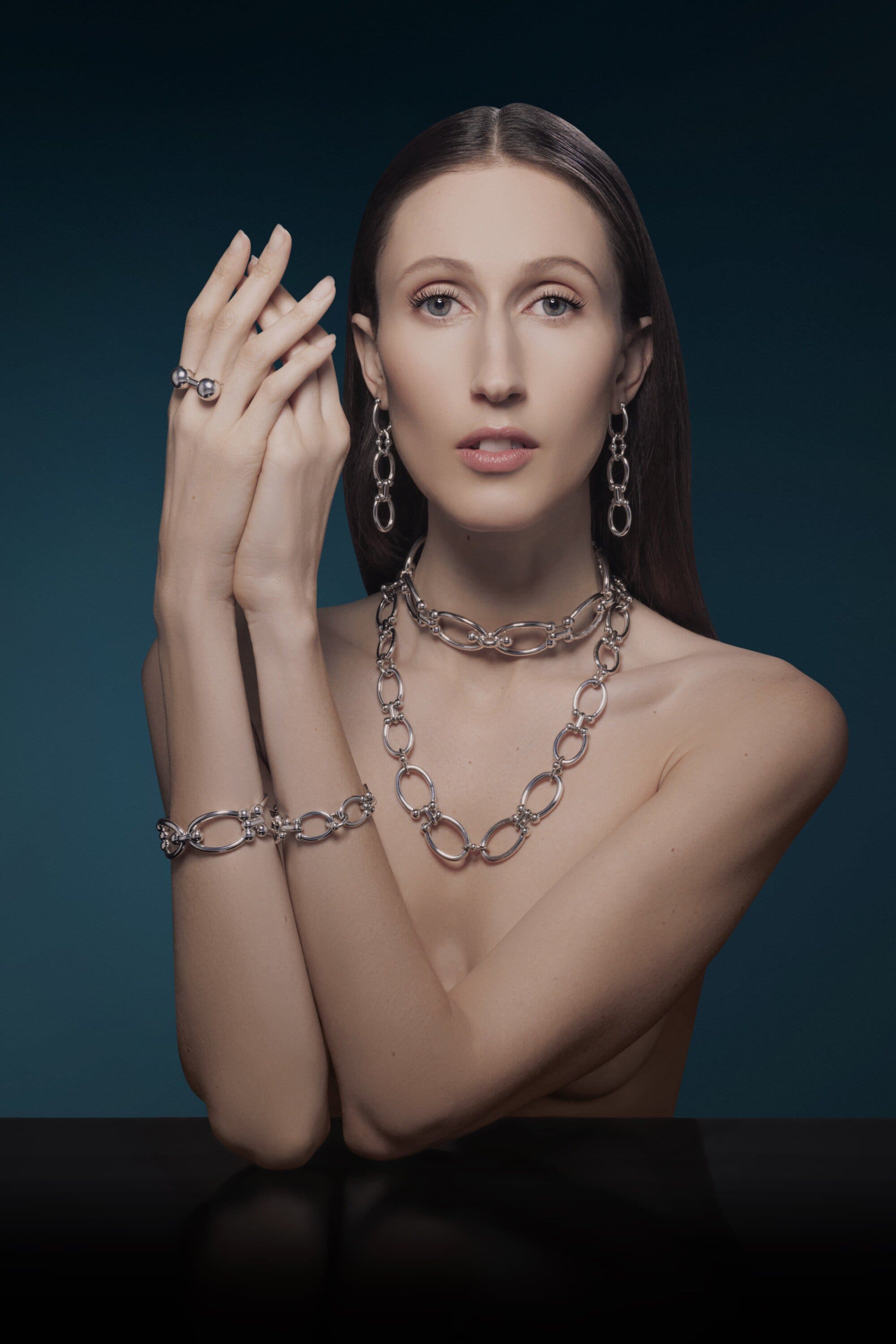 Close-up shot of a woman wearing silver statement jewelry including a necklace, bracelet, ring, and drop earrings on a dark background.