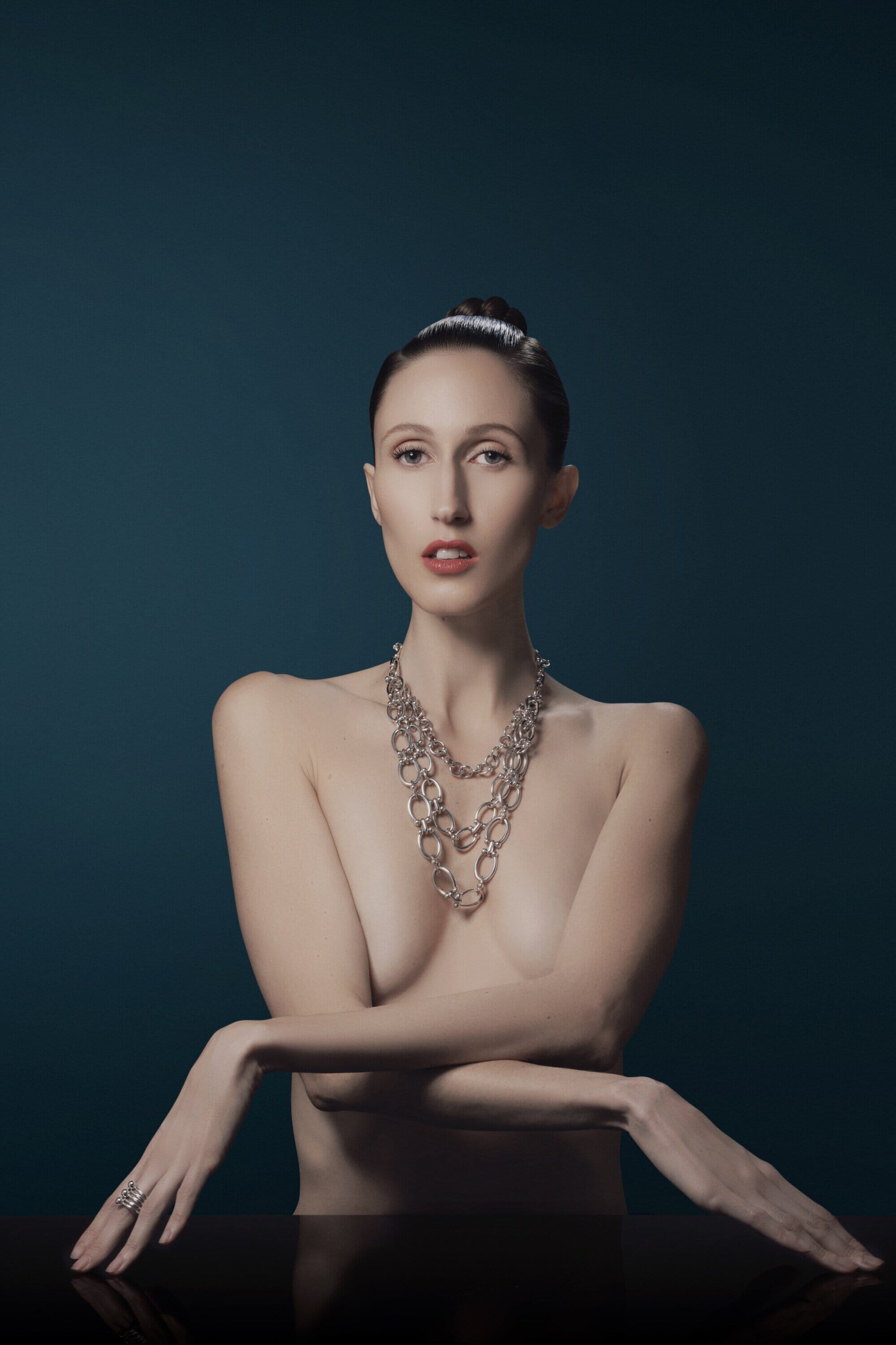 Elegant woman with a silver necklace poses in a studio with a teal background, her arms crossed over a shiny black table.