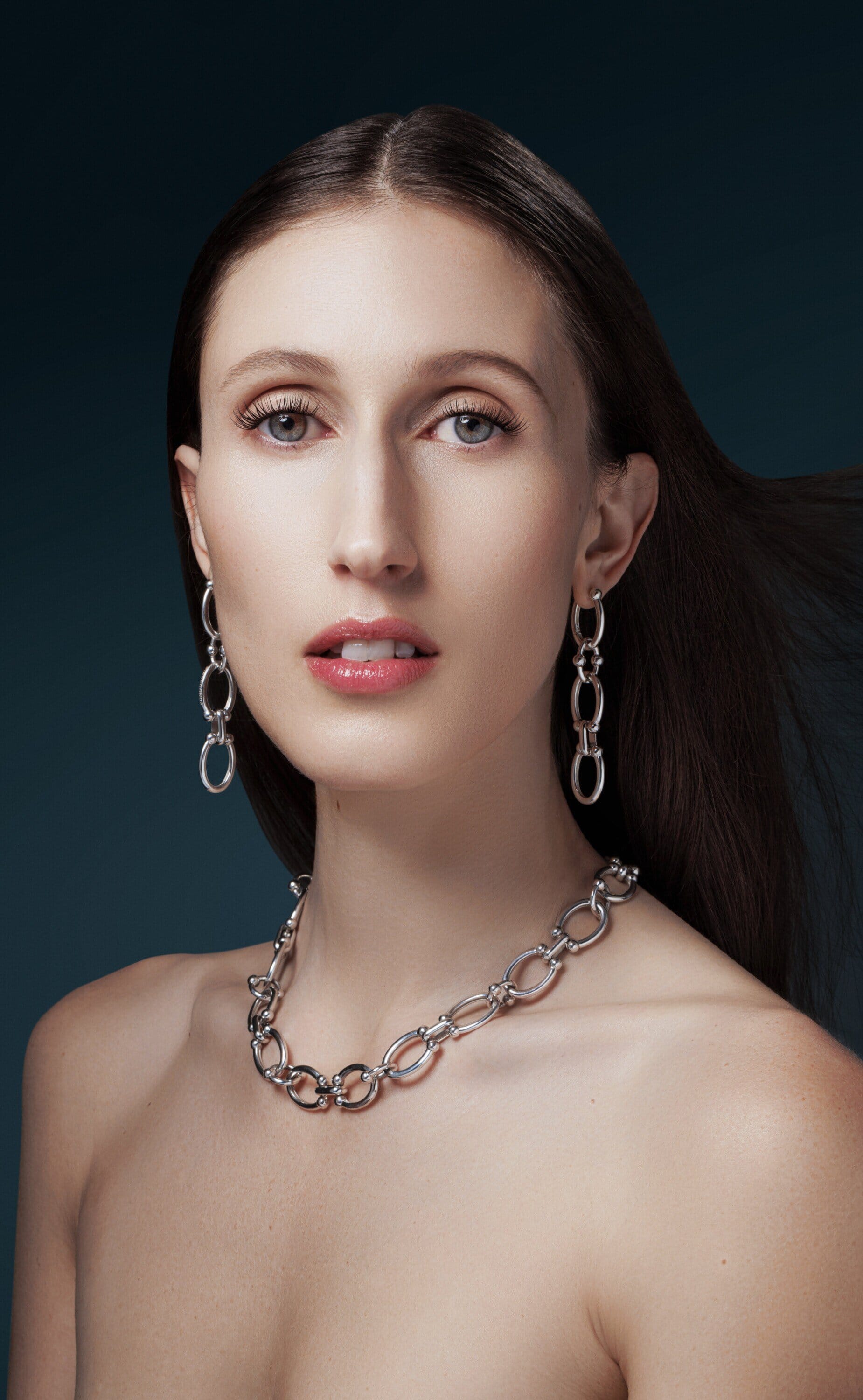 Close-up studio portrait of a young adult woman with long brown hair wearing silver earrings and a necklace against a dark teal backdrop.