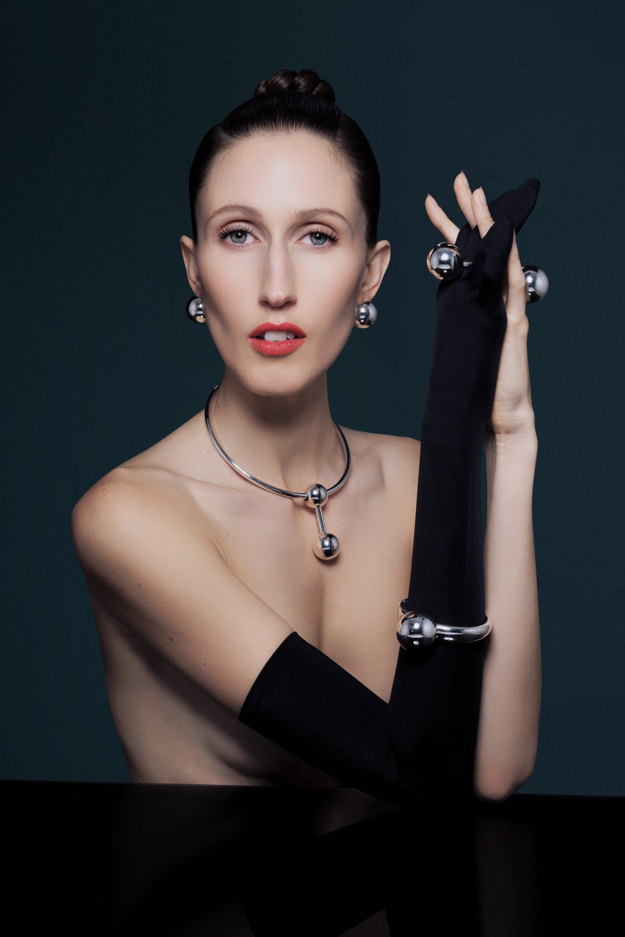 Elegant woman wearing silver jewellery and black gloves poses in a studio with red lipstick and a dark background, looking at the camera.