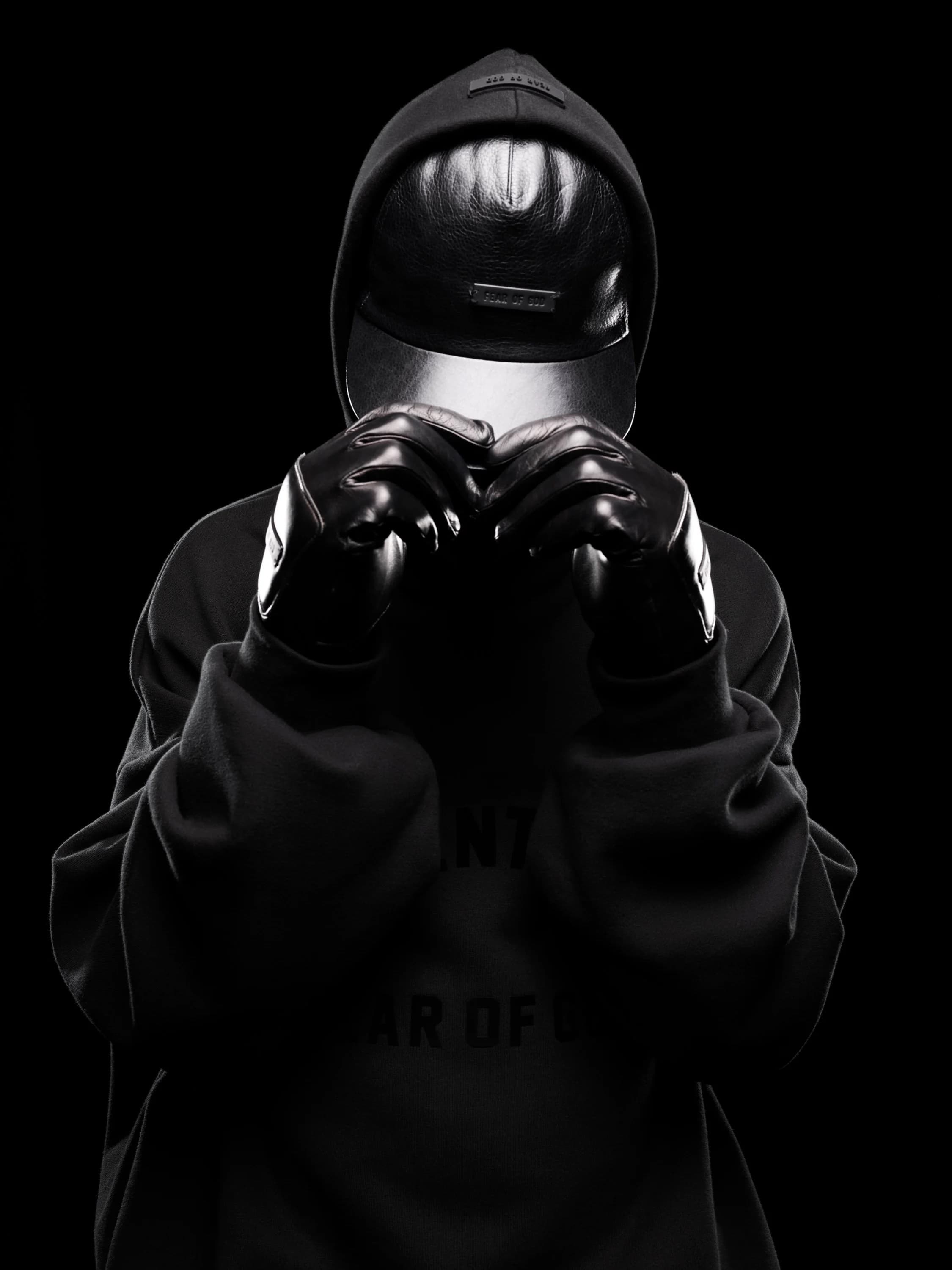 Person in a black hoodie and mask with black gloves against a dark background, creating a mysterious and obscured figure in shadow.