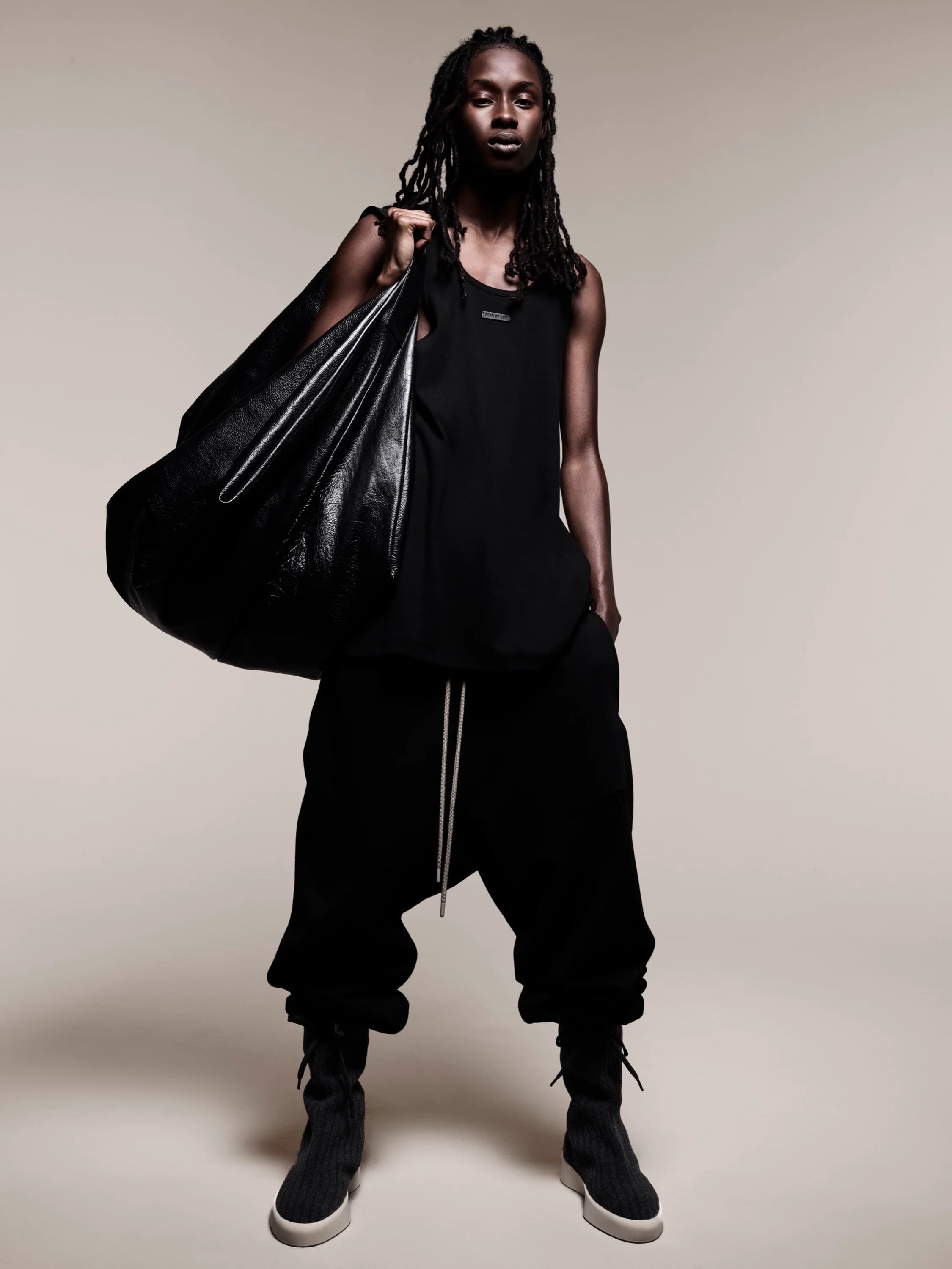 Full-length studio shot of a Black adult male model in a dark outfit carrying a large black bag over his shoulder in a neutral studio.