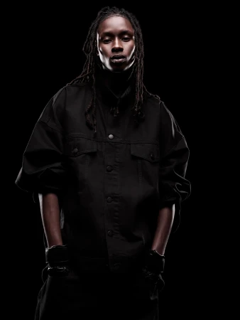 Dark-skinned adult with dreadlocks poses in a black jacket and turtleneck against a black backdrop in a dramatic studio portrait.