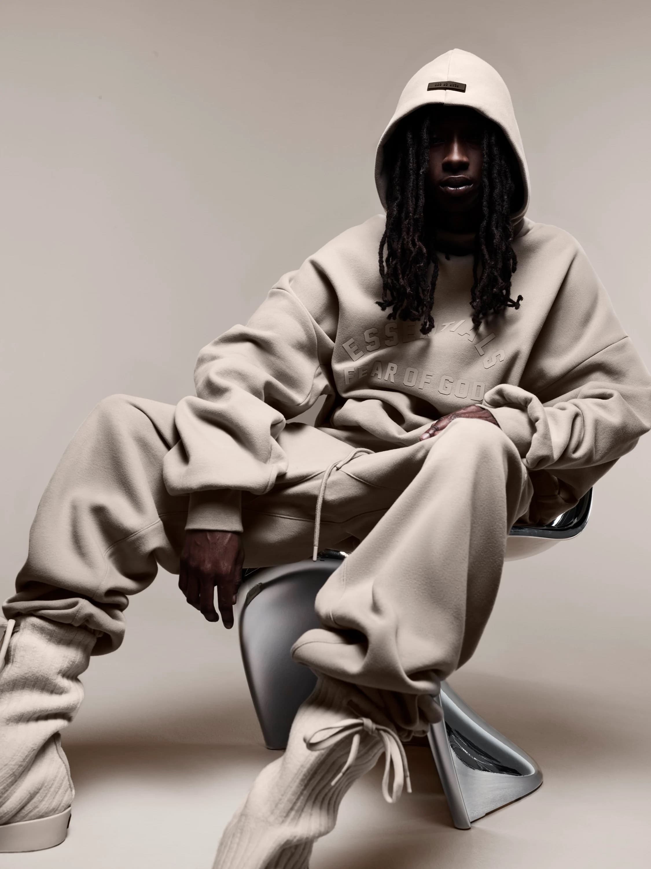 Man in a beige hoodie and sweatpants sits on a chair in a studio setting, showcasing casual streetwear fashion.