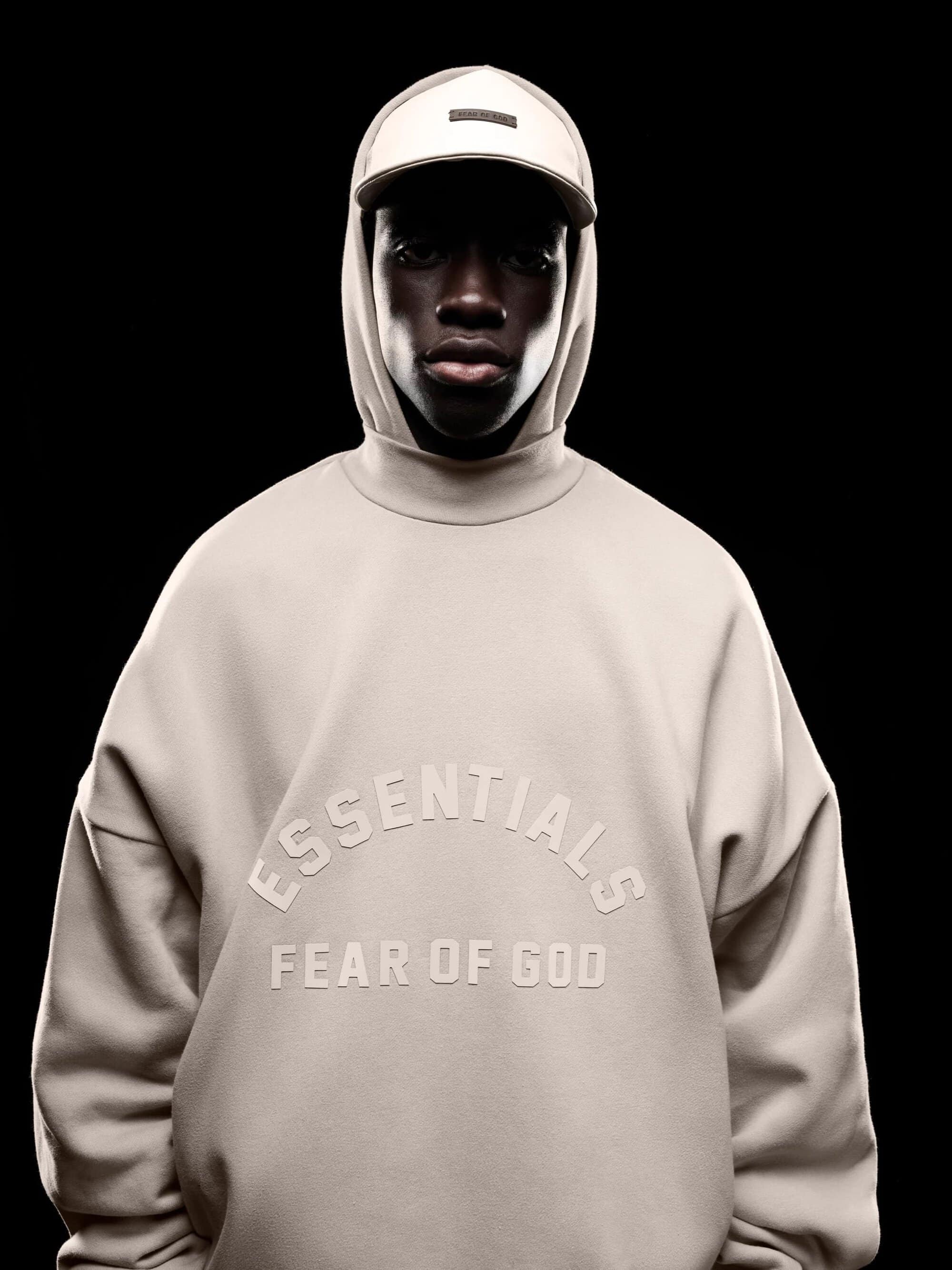 Portrait of African American male model in a studio wearing a beige hoodie with ESSENTIALS logo and baseball cap against a black background.