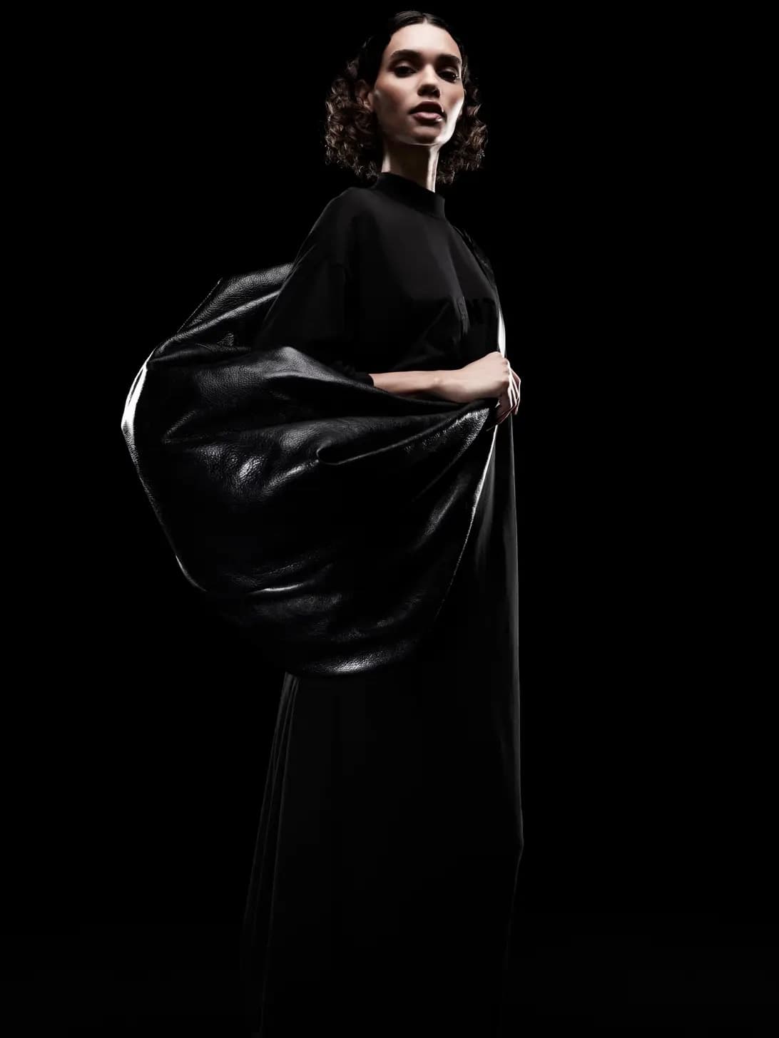A woman in a black dress and draped fabric poses in a dark studio, illuminated by dramatic lighting, showcasing fashion design and style.