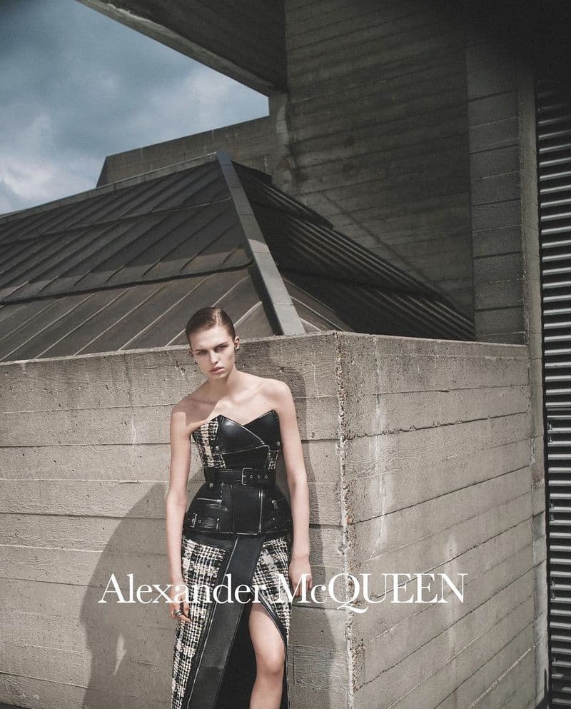 Woman poses in Alexander McQueen dress by a concrete structure, under a cloudy sky, in editorial fashion photography. 