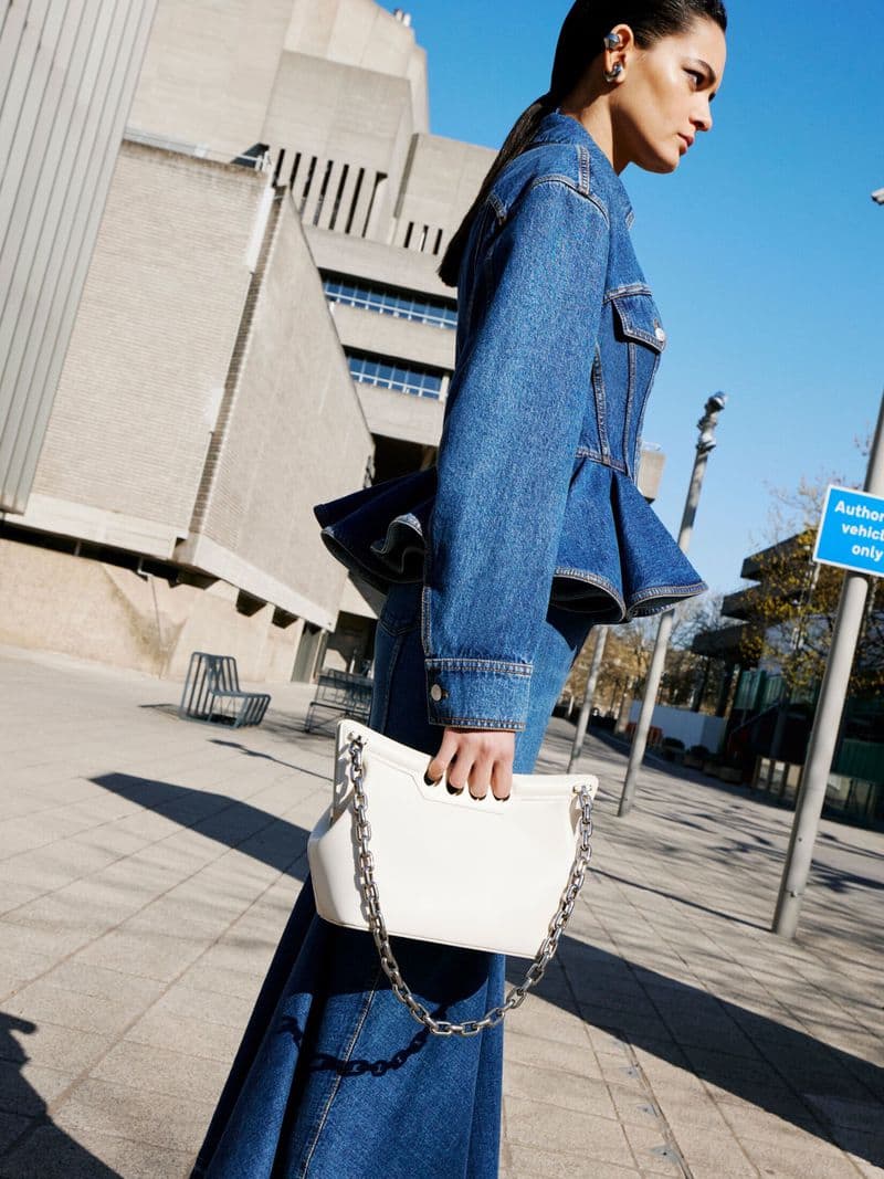 Model in dark denim peplum jacket holding a white chain handbag against brutalist concrete architecture.