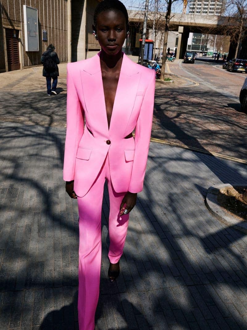 A slender Black woman in a vibrant pink suit walks down a city sidewalk, with sunlight casting shadows on the paved surface.