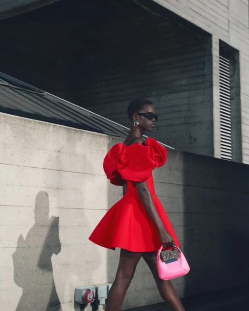 A dark-skinned female model wears a red dress with puffy sleeves and carries a pink purse, posing on a city street near concrete walls.