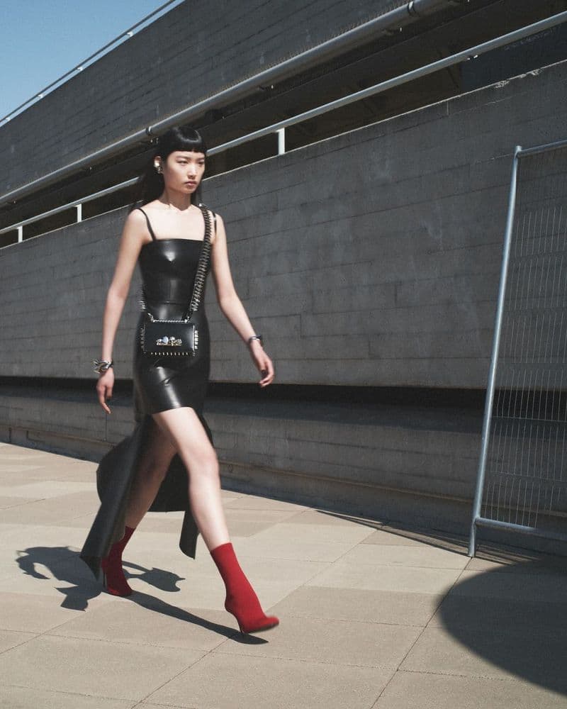 A woman in a black dress and red socks walks on a paved surface in daylight, with an urban concrete building in the background.