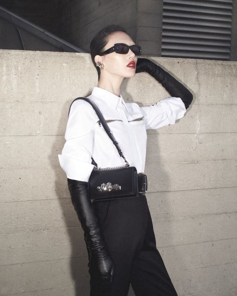 Model in white shirt, black trousers, long leather gloves, and sunglasses standing against a concrete wall