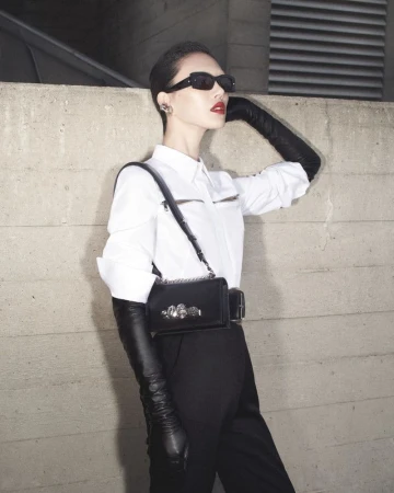 Model in white shirt, black trousers, long leather gloves, and sunglasses standing against a concrete wall