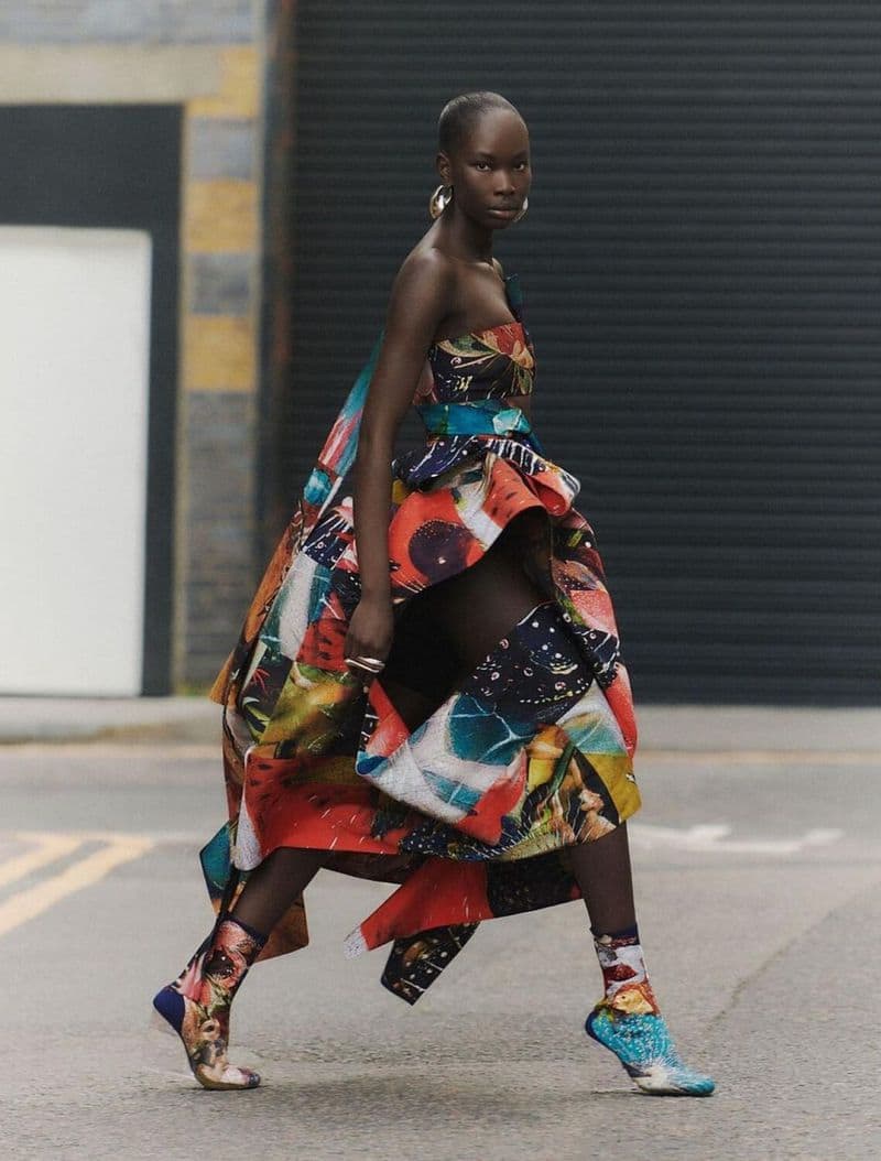 A stylish dark-skinned woman in a strapless, multicolored high-low dress and art-printed sock boots walks on a city street.