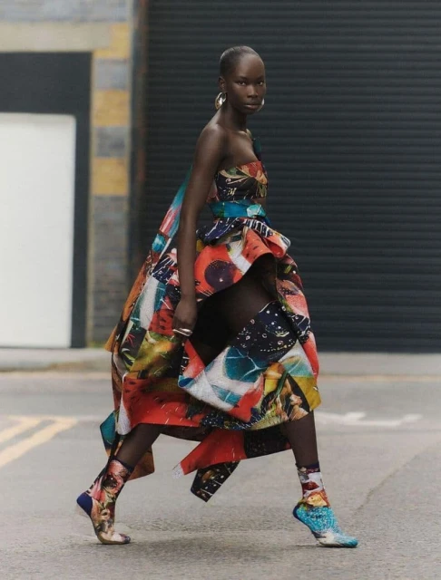 A stylish dark-skinned woman in a strapless, multicolored high-low dress and art-printed sock boots walks on a city street.