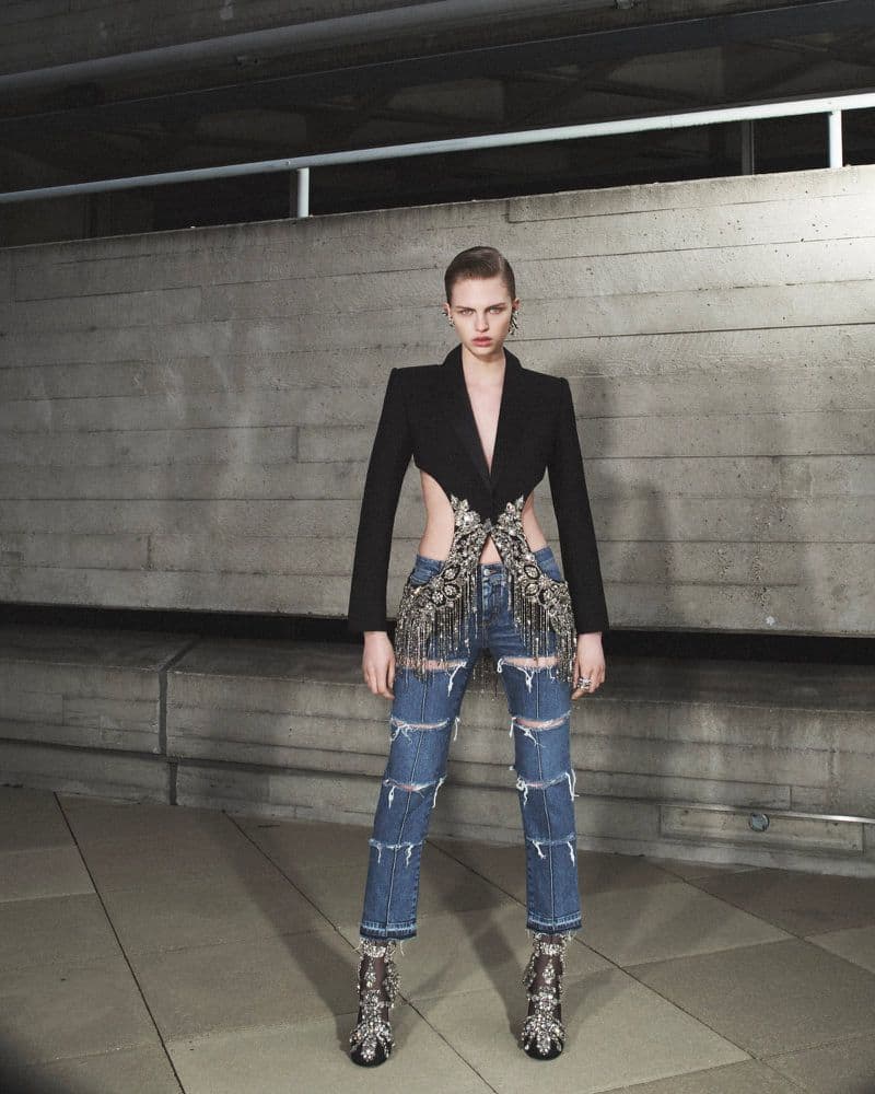 Fashion model poses in a black blazer, distressed denim, and embellished heels against a concrete wall in a medium shot.